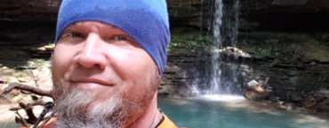 A man wearing a blue beanie and organge shirt is standing in front of a blue waterfall.